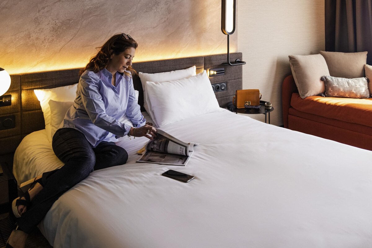A person sitting on a bed in a hotel room, reading a magazine.