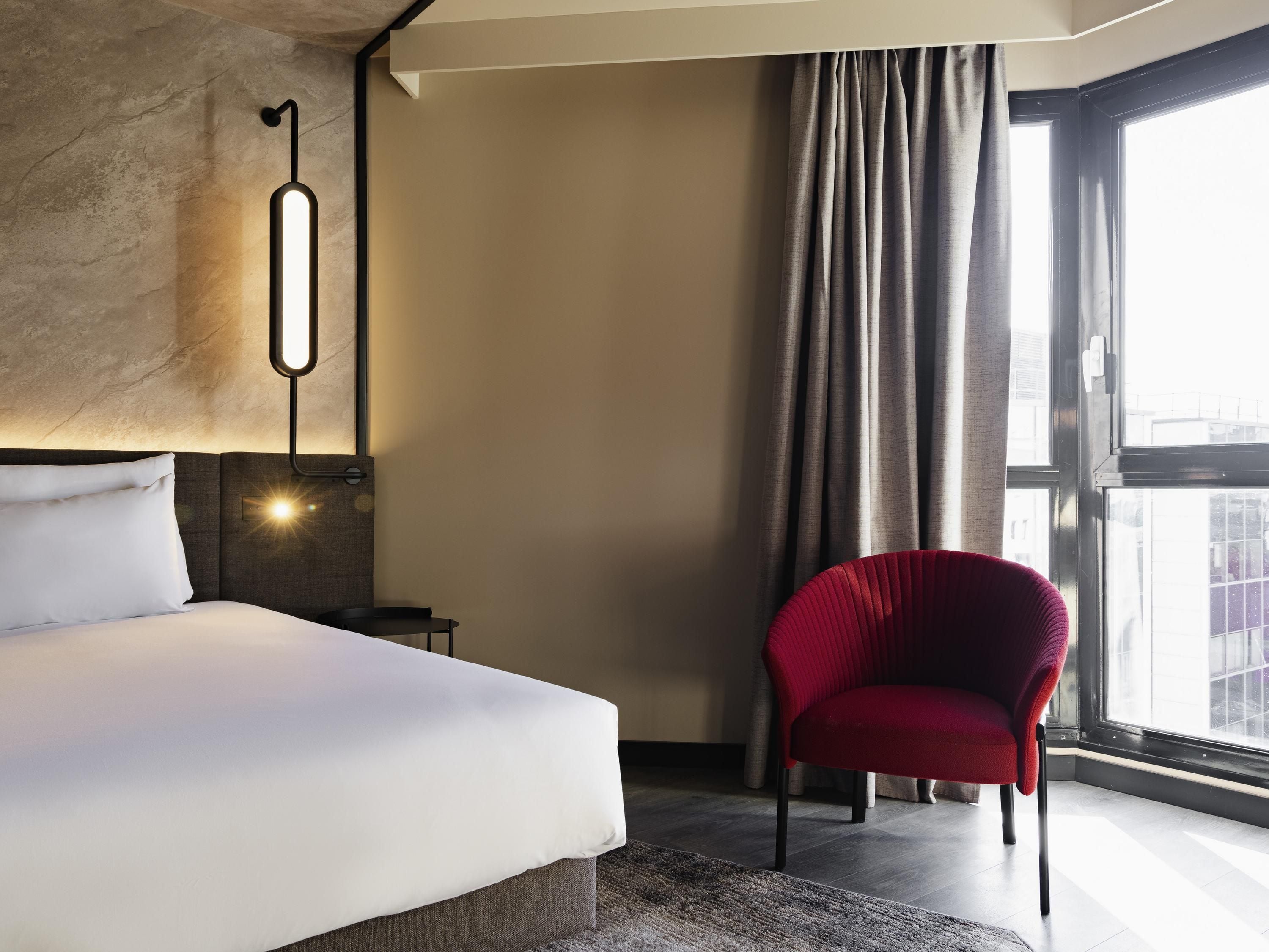 Habitación de hotel con cama, una silla roja, una lámpara de pared y un ventanal con cortinas.