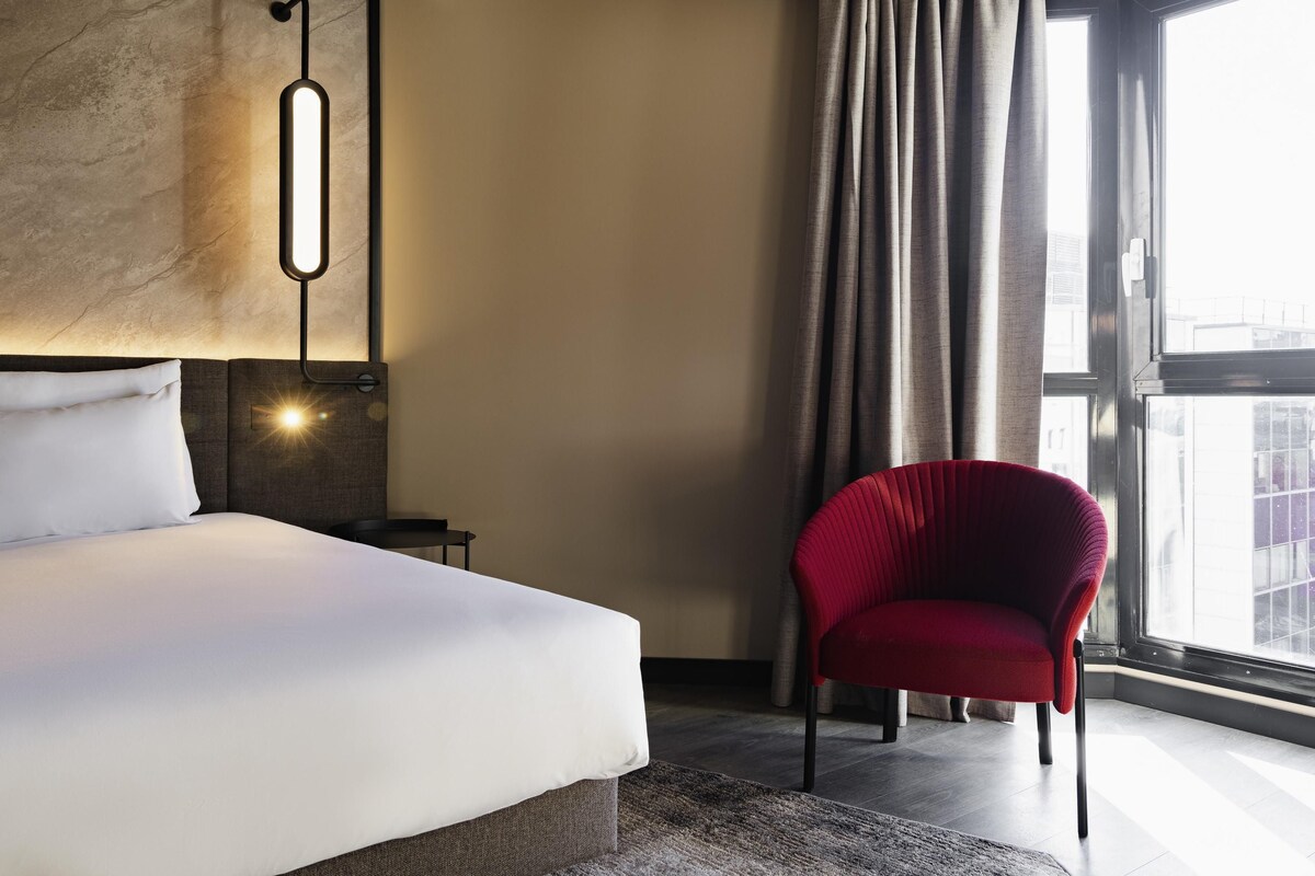 A hotel room with a bed, a red chair, a wall-mounted light, and a large window with curtains.
