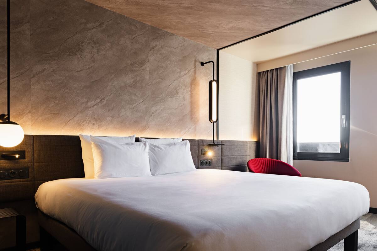 Ein modernes Hotelzimmer mit einem großen Bett, einem Kopfteil mit integrierter Beleuchtung, einem Nachttisch mit Lampe und einem Vorhangfenster.