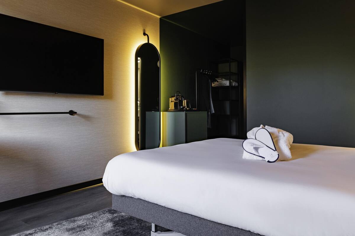 Ein modernes Hotelzimmer mit einem großen Bett, einem flachbildfernseher an der Wand und einem Nachttisch mit Lampe.