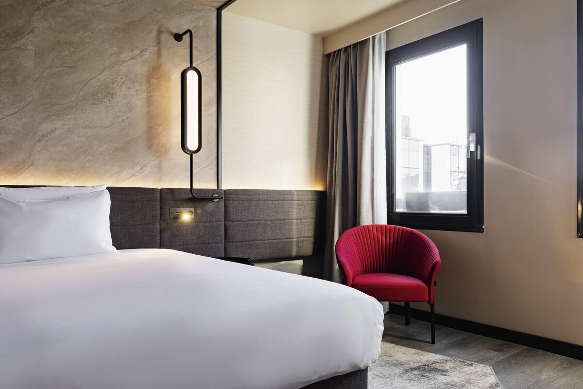 Ein modernes Hotelzimmer mit einem großen Bett, einem roten Sessel, einem großen Fenster mit Vorhängen und einer Wandleuchte.