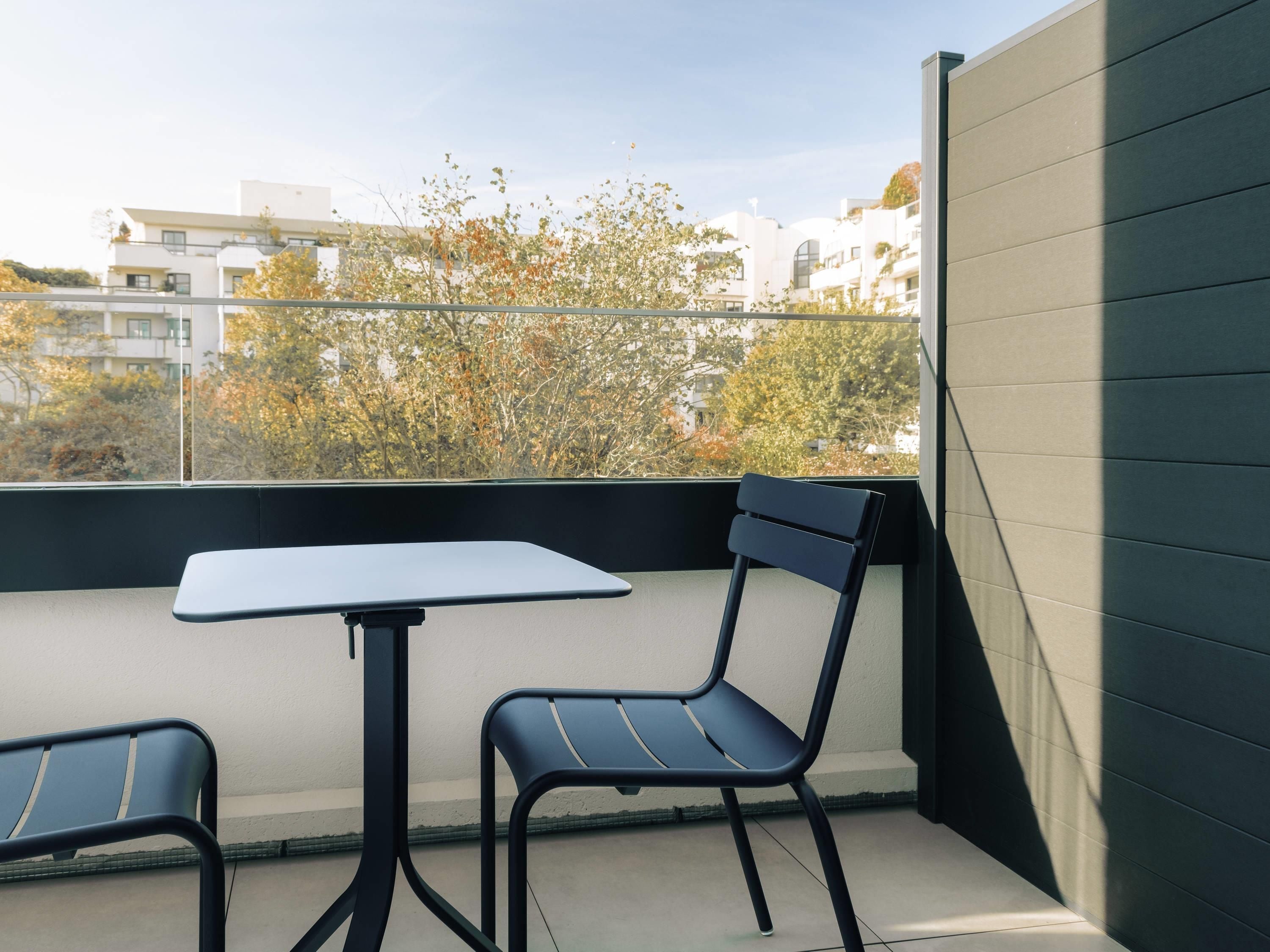 Uma varanda com mesa e duas cadeiras, vista para edifícios e árvores, e uma parede de cor escura.