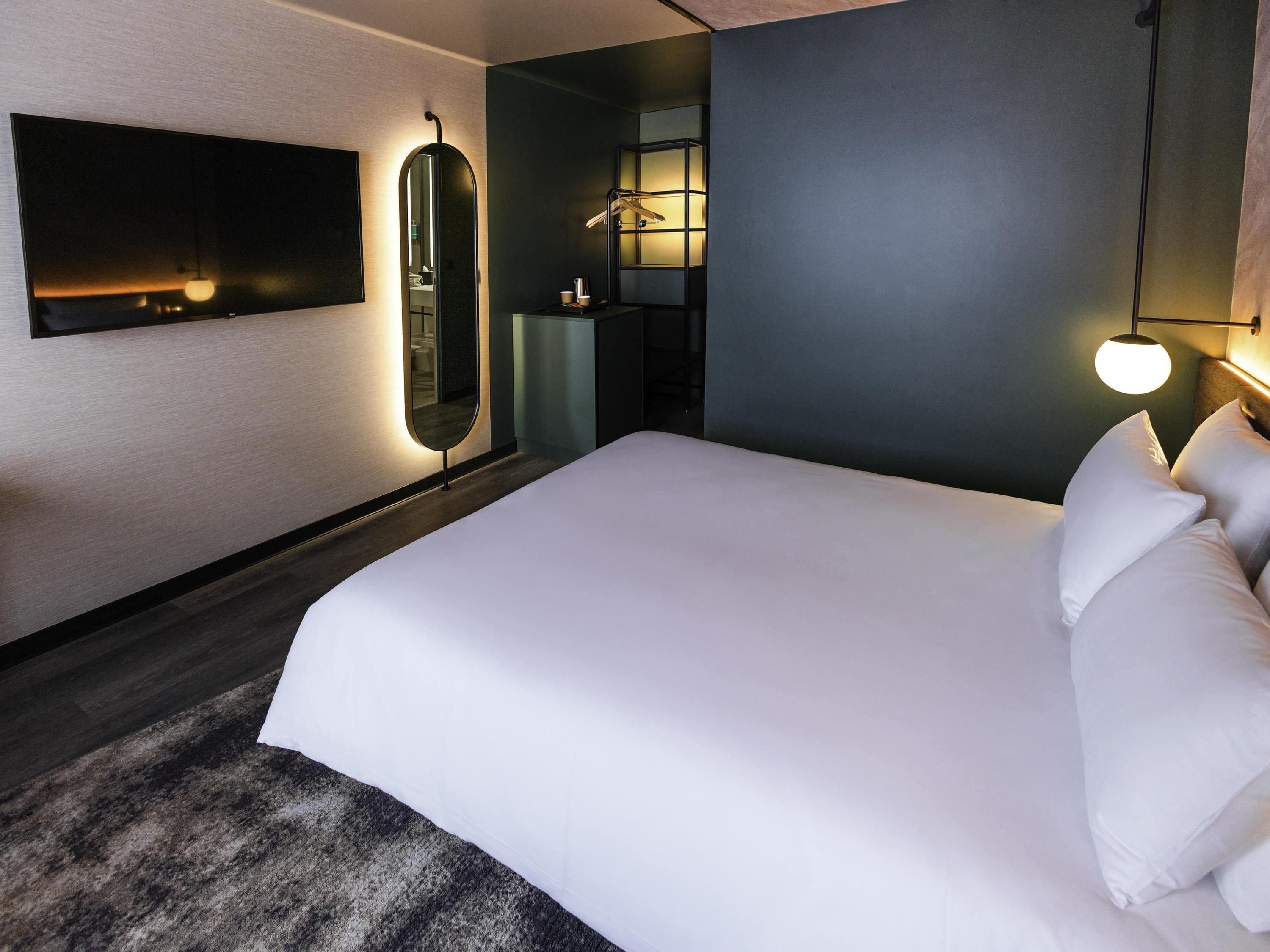 A modern hotel room with a large bed, a wall-mounted TV, a mirror, and a bedside lamp.