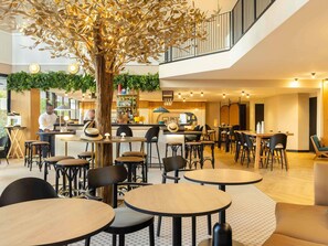 A modern indoor dining area with round tables and chairs, a central tree, and a bar area with a counter. - Novotel Paris Suresnes Longchamp (Suresnes)