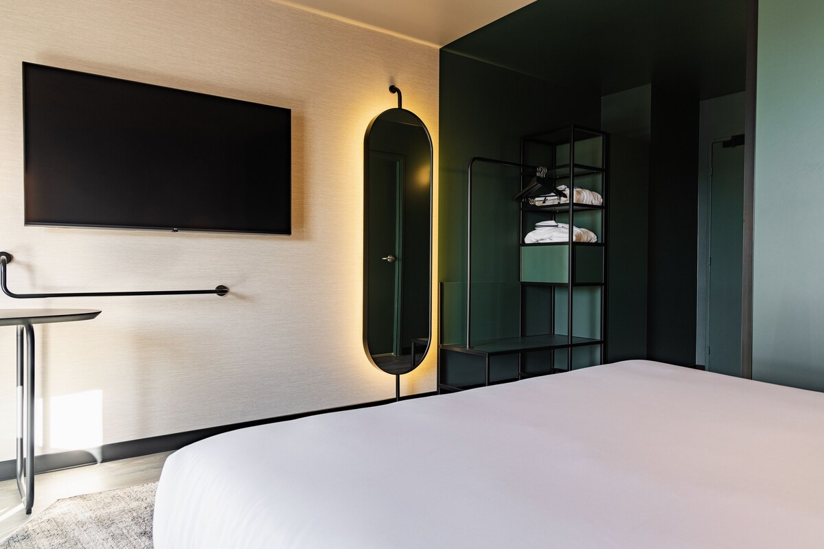 A modern hotel room with a large bed, a flat-screen TV mounted on the wall, a bedside table, and a built-in wardrobe with shelves.