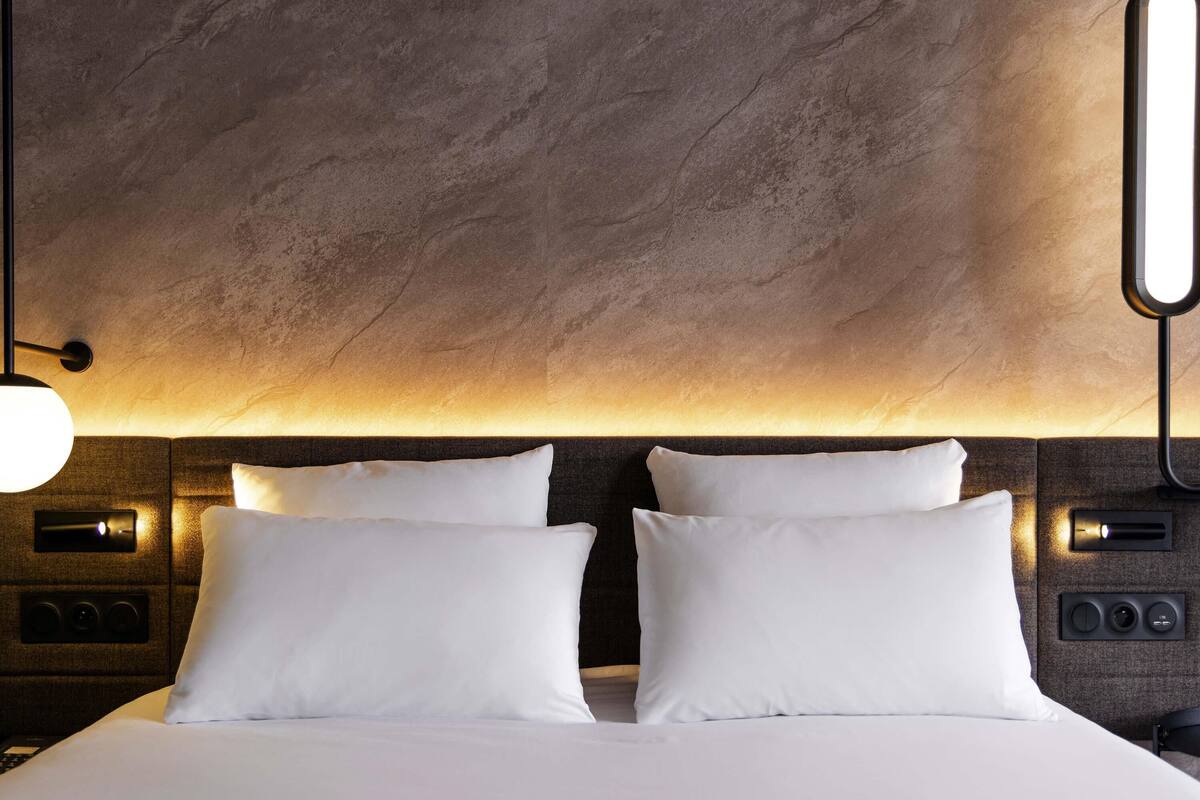 Ein modernes Hotelzimmer mit einem großen Bett, zwei weißen Kissen, einer Kopfteilbeleuchtung und einer wandmontierten Bedienoberfläche.