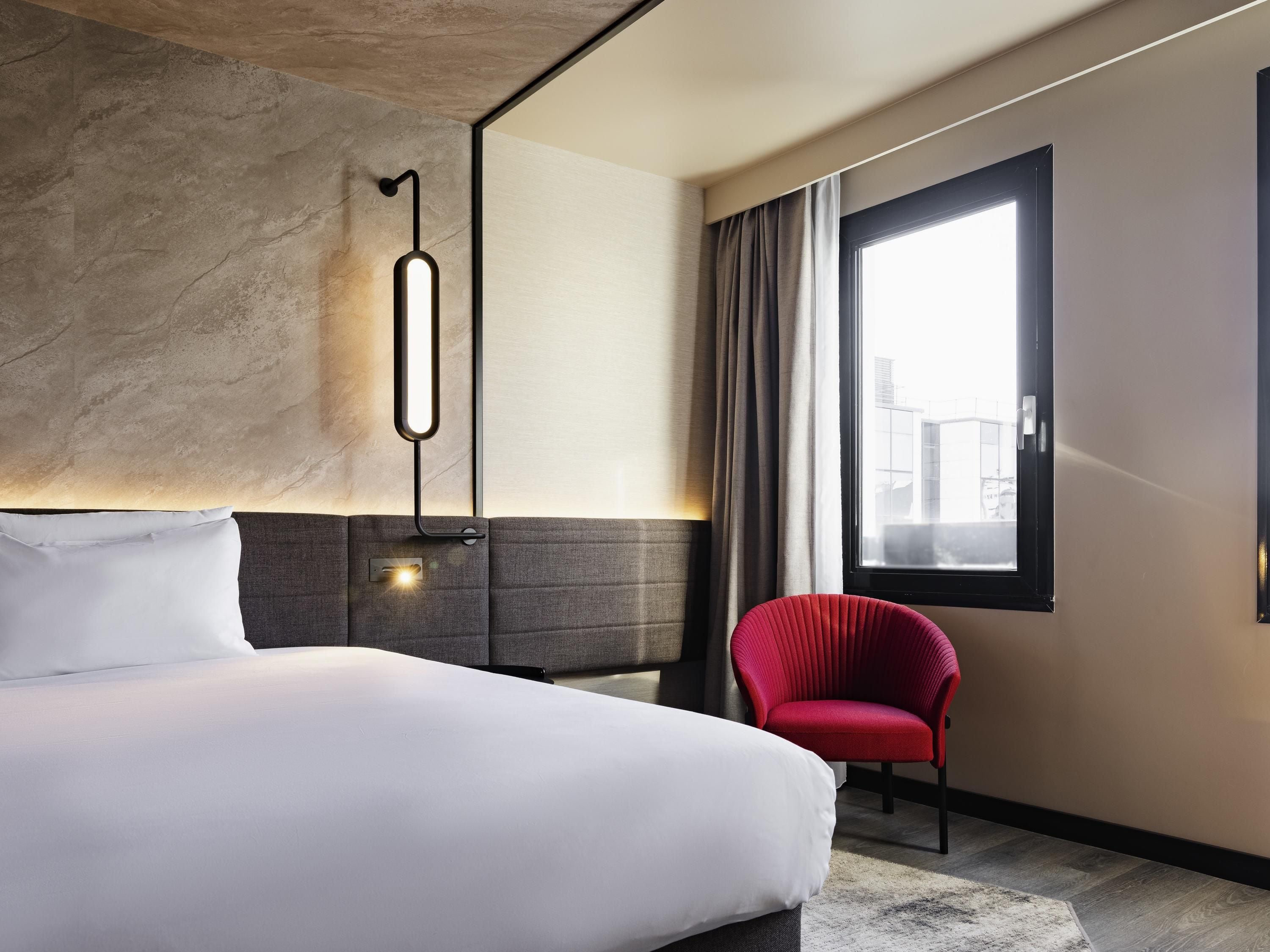 A modern hotel room with a large bed, a red chair, a large window with curtains, and a wall-mounted light fixture.