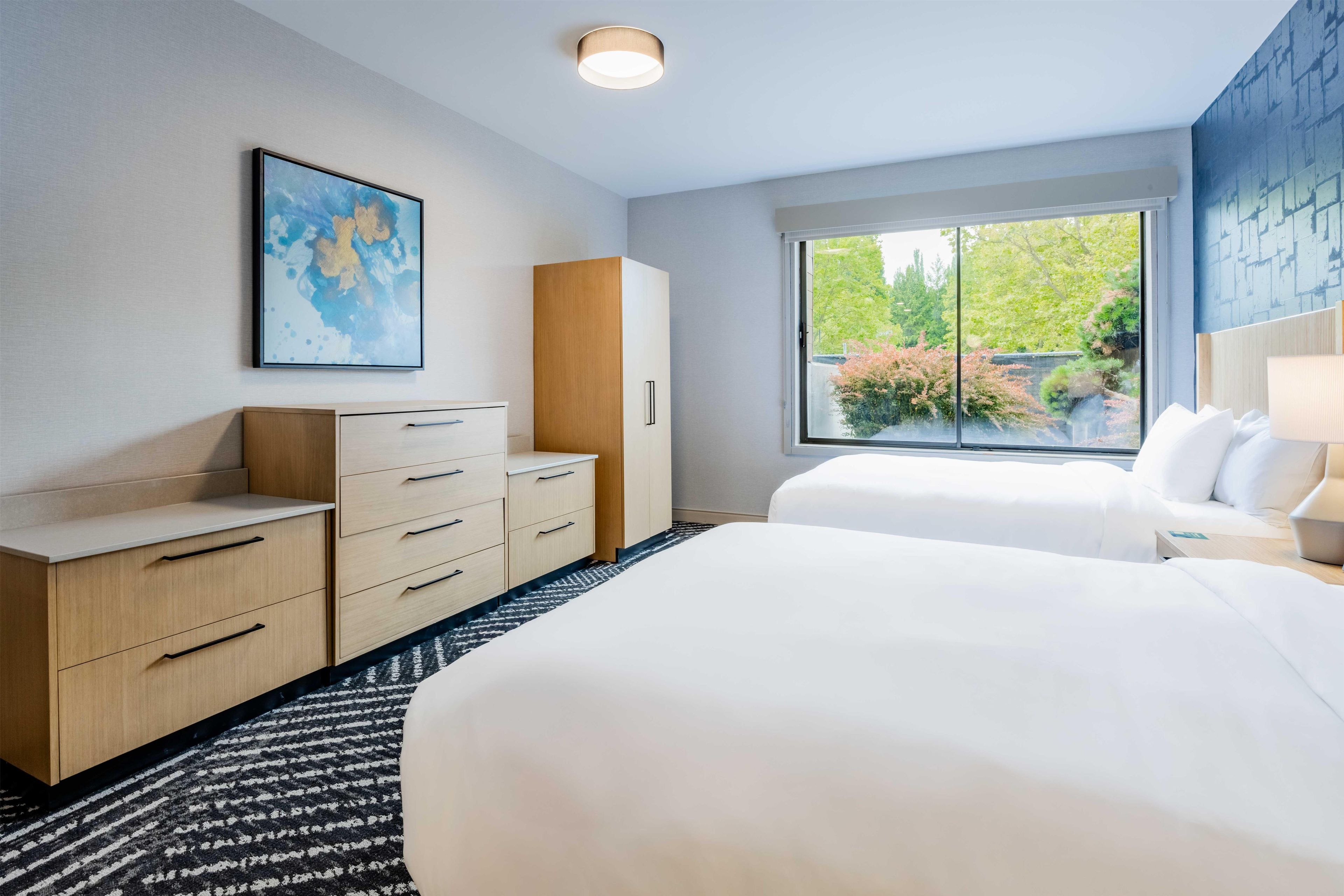 A hotel room with a bed, a desk, a wardrobe, a window with a view of greenery, and a painting on the wall.