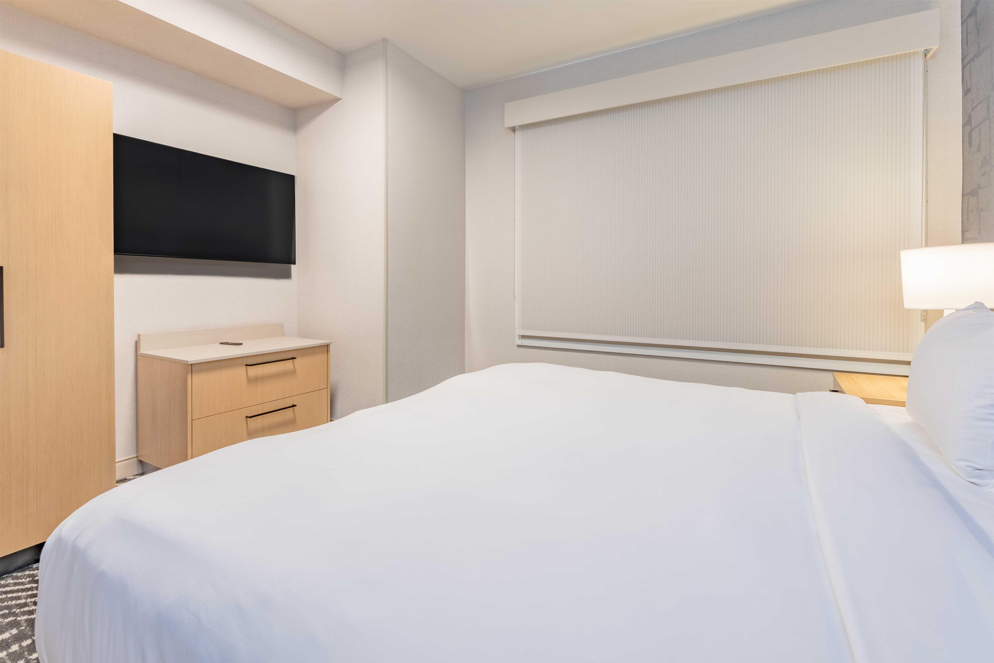 Hotelkamer met een bed, een aan de muur bevestigde televisie, een nachtkastje en een raam met zonwering.