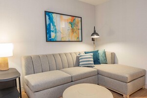 Un salon moderne avec un canapé d’angle, une table basse ronde, un lampadaire et un tableau abstrait encadré accroché au mur.