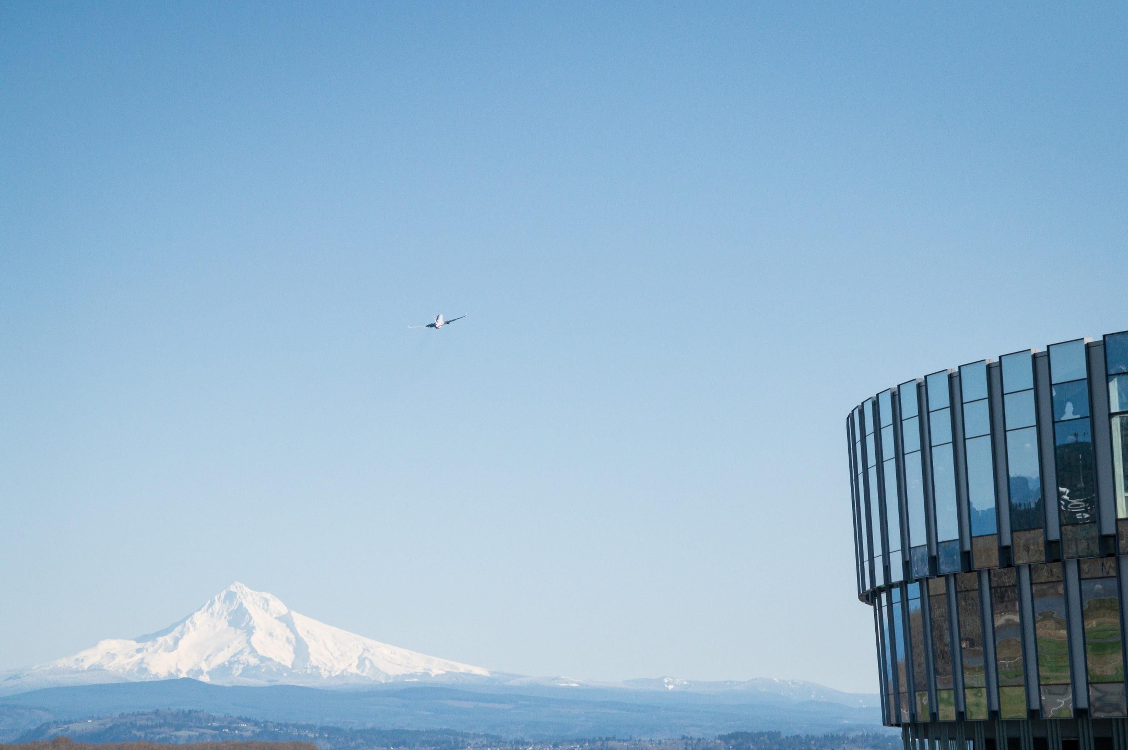 A snow-capped mountain, a modern building with reflective glass windows, and a plane in the sky.
