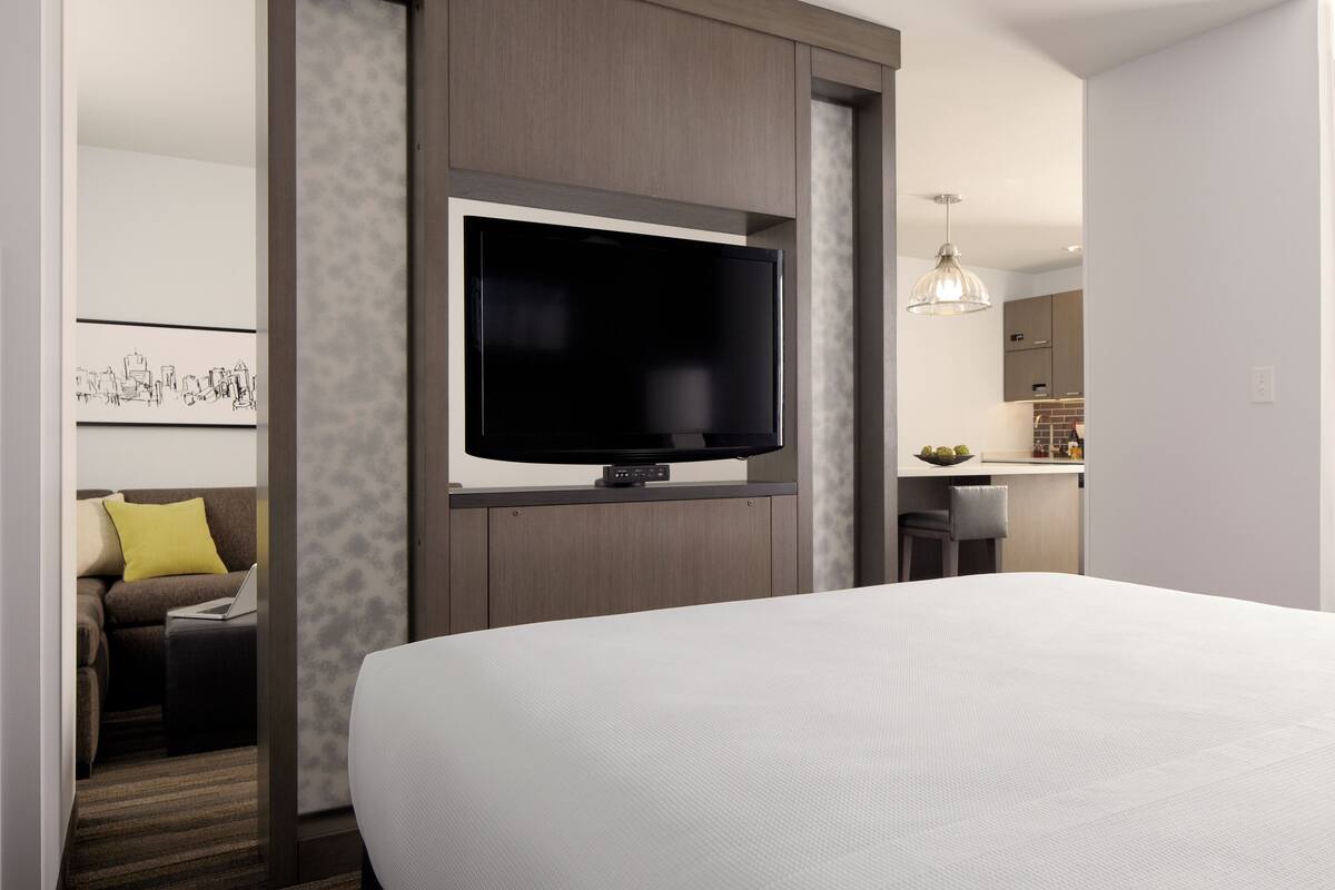 A modern hotel room with a large bed, a flat-screen TV mounted on the wall, a small seating area with a sofa and ottoman, and a kitchenette in the background.