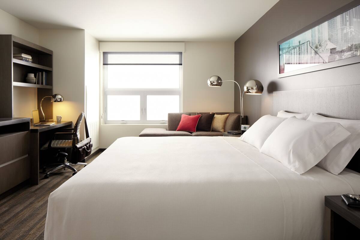 A hotel room with a large bed, a desk with a chair, a bookshelf, and a window providing natural light.