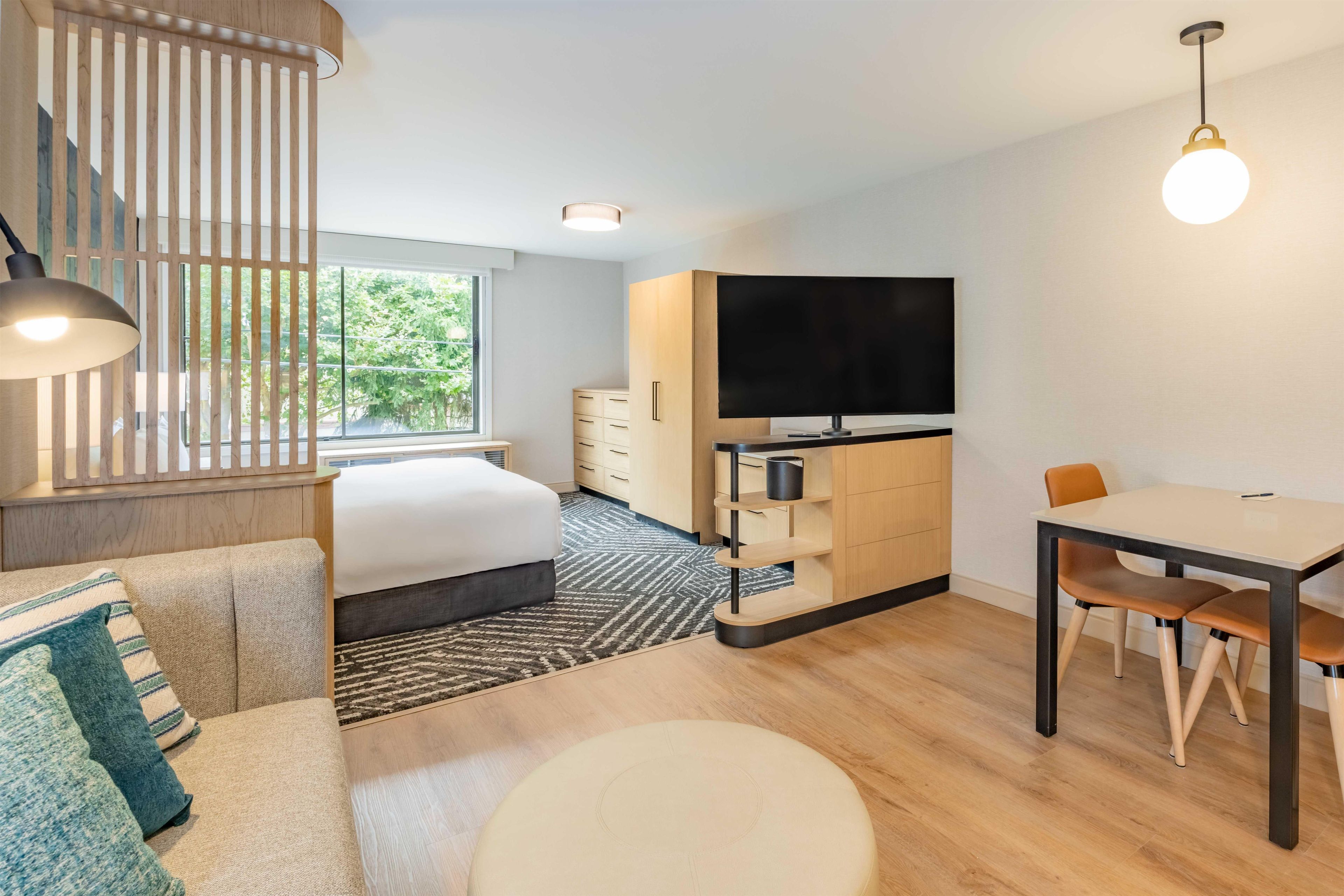 A modern hotel room with a bed, a desk, a TV, and a dining area.