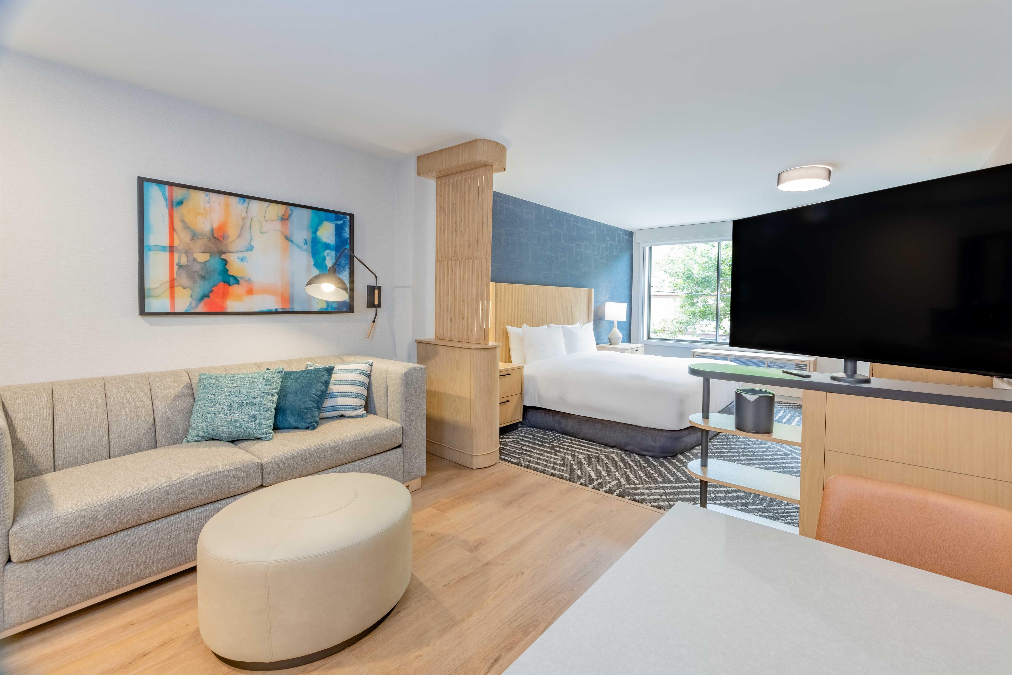 Una habitación de hotel moderna con sofá, reposapiés, cama, televisión y una obra de arte en la pared.