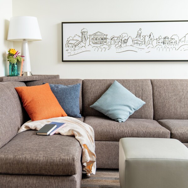 A living room with a grey sofa, two pillows, a blanket, and a book. A framed picture hangs on the wall above the sofa.