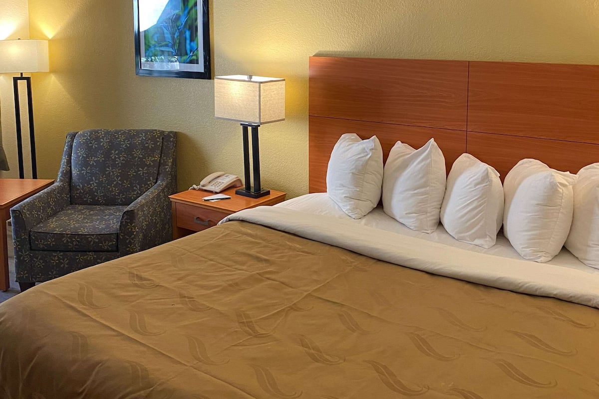 A hotel room with a large bed, a wooden headboard, a chair, a nightstand with a lamp, and a painting on the wall.