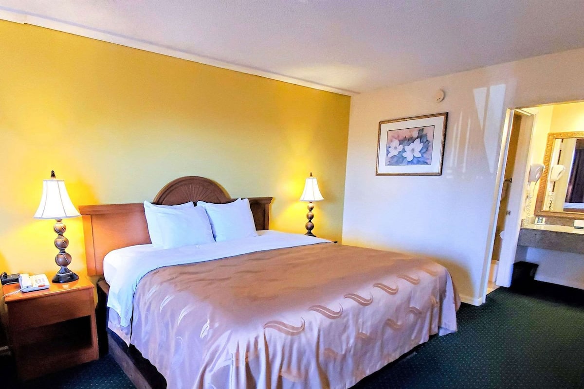 A hotel room with a bed, two bedside lamps, a nightstand, a painting on the wall, and a bathroom mirror.