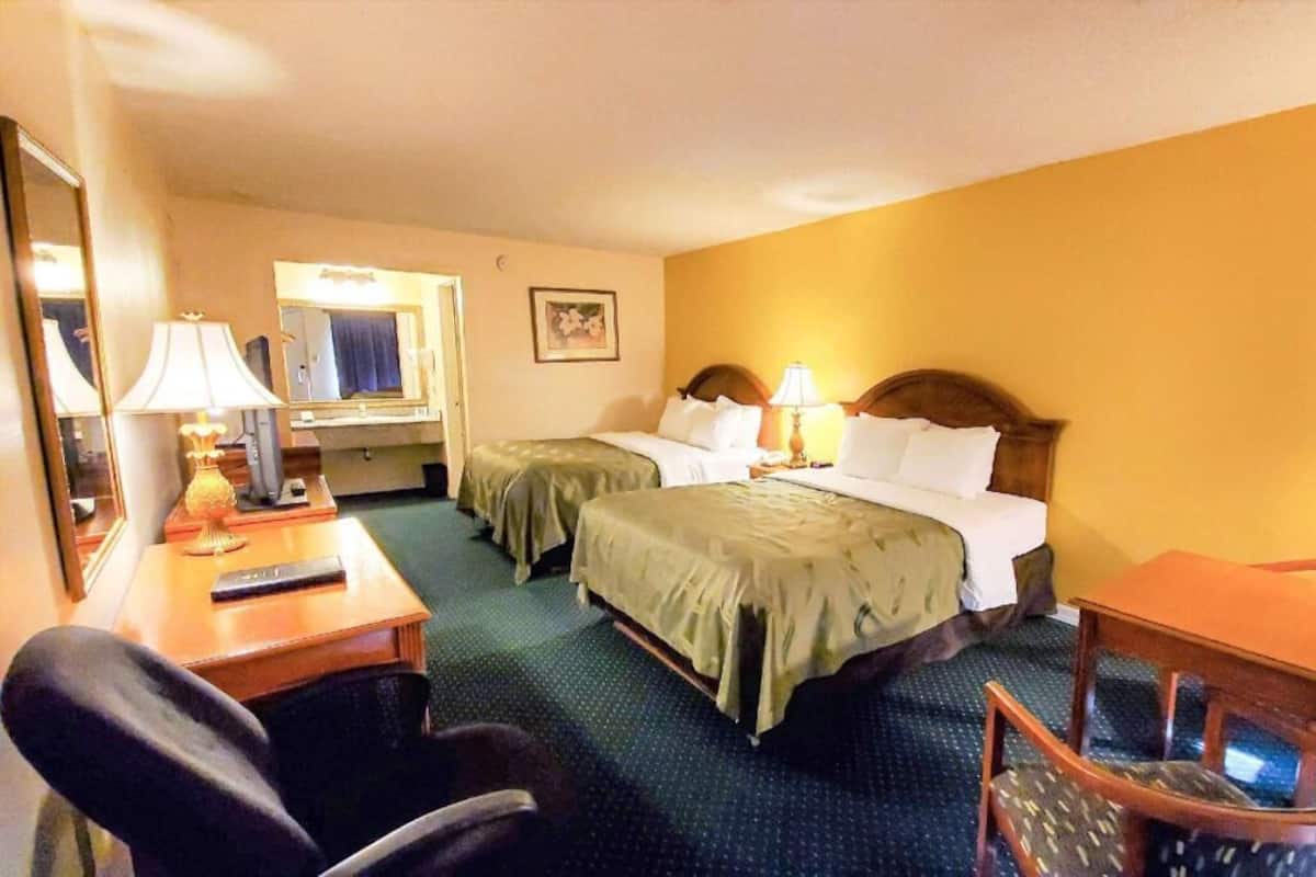 A hotel room with two beds, a desk, a chair, a mirror, and a lamp.