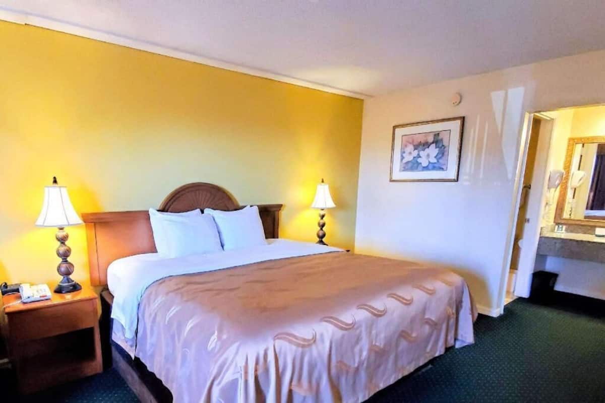A hotel room with a bed, two bedside lamps, a nightstand, a framed picture on the wall, and a bathroom visible through an open door.