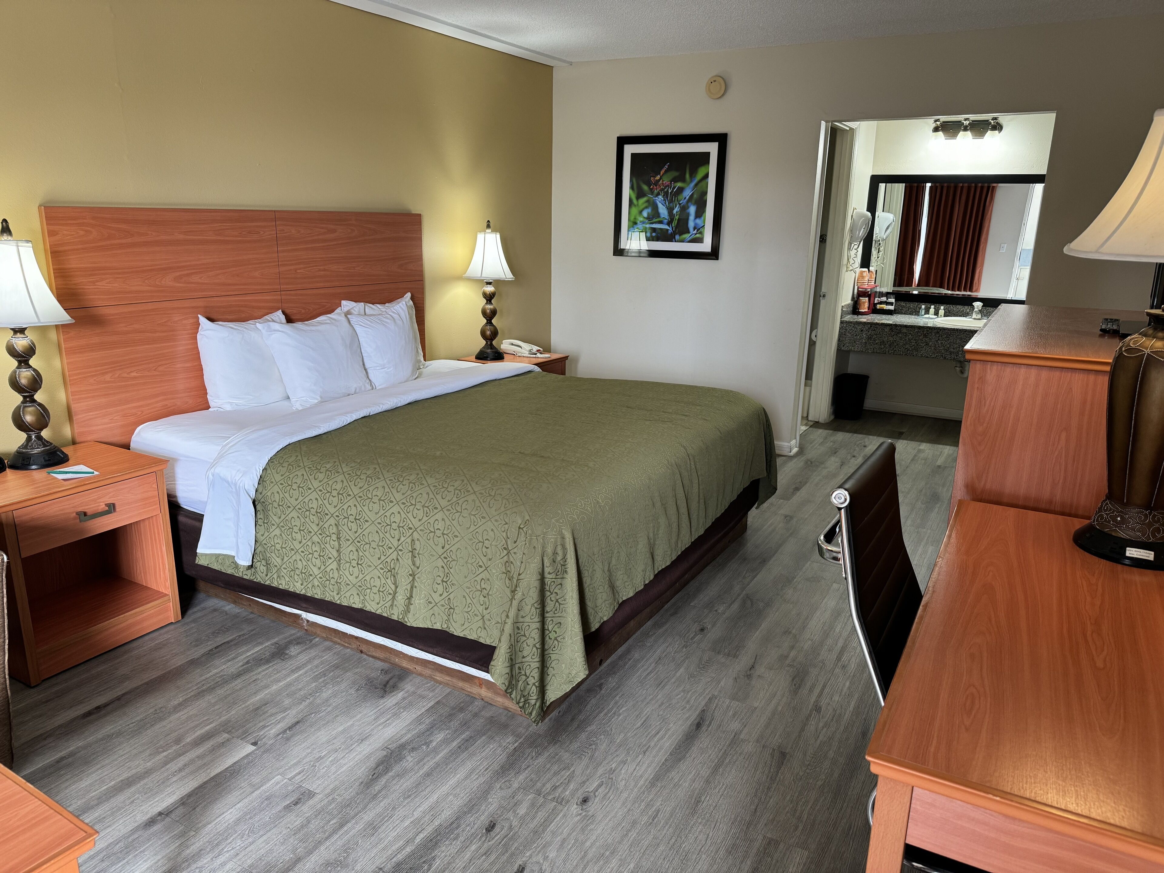Habitación de hotel con cama de cabecera de madera, cubrecama verde, mesita de noche con lámpara, escritorio con silla y baño visible a través de una puerta abierta.