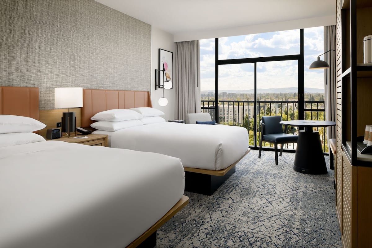 A hotel room with two beds, a desk, a chair, a small table, and a view of the city through large windows.