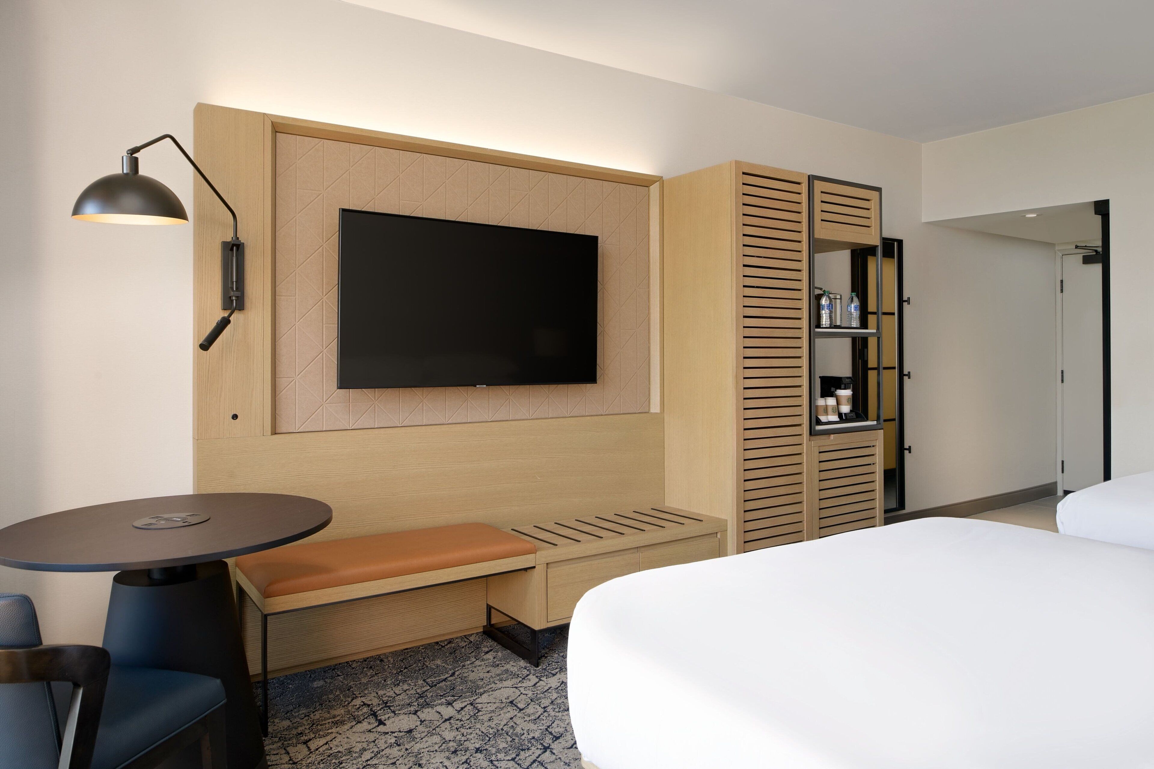 A hotel room with a bed, a desk with a chair, a TV mounted on the wall, a shelf with bottles, and a wardrobe with sliding doors.