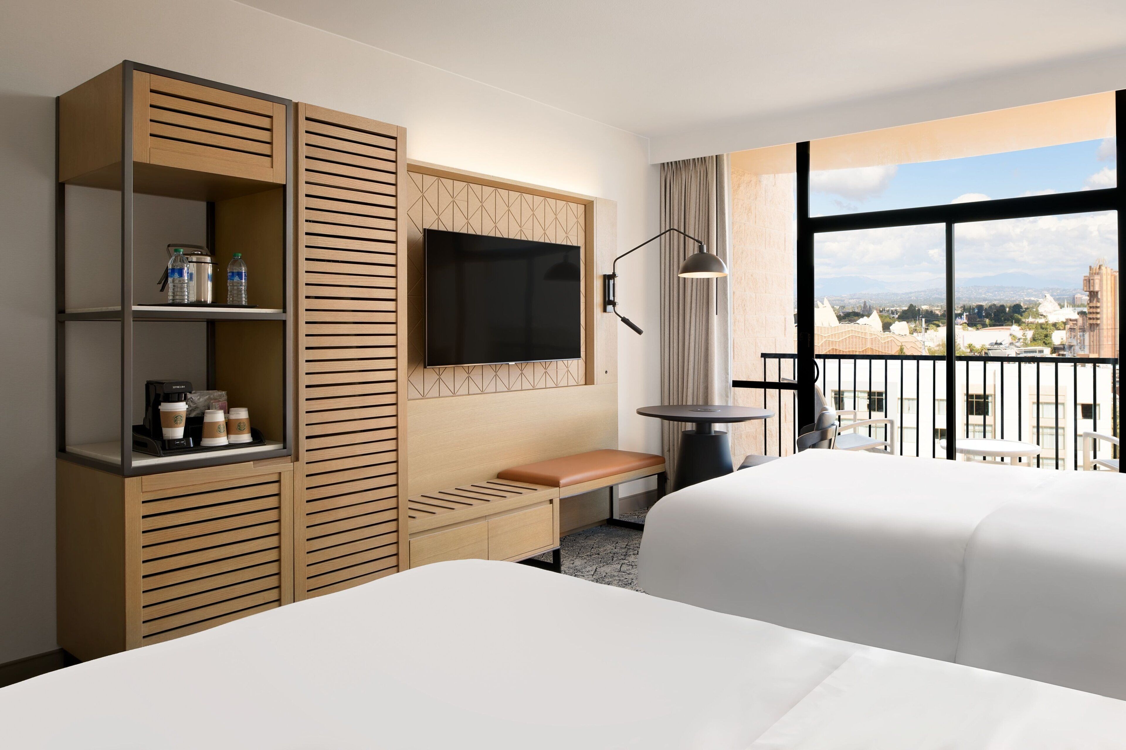 A hotel room with a large bed, a TV mounted on the wall, a wooden cabinet with shelves, a small table, and a balcony with a view of the city.