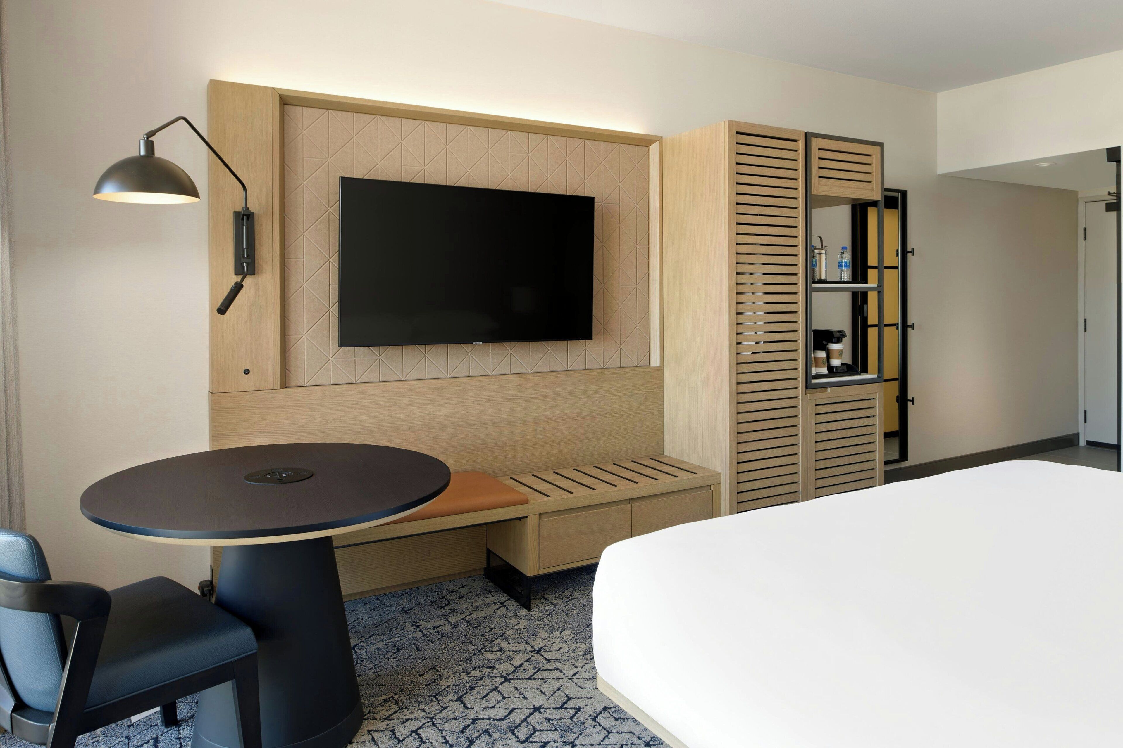 Ein Hotelzimmer mit Bett, einem an der Wand montierten Fernseher, einer Sitzecke mit Tisch und Stühlen sowie einem Kleiderschrank mit Spiegel.