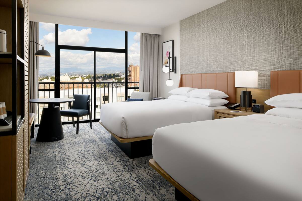 A hotel room with two beds, a desk, and a chair. There is a large window with a view of buildings and mountains.
