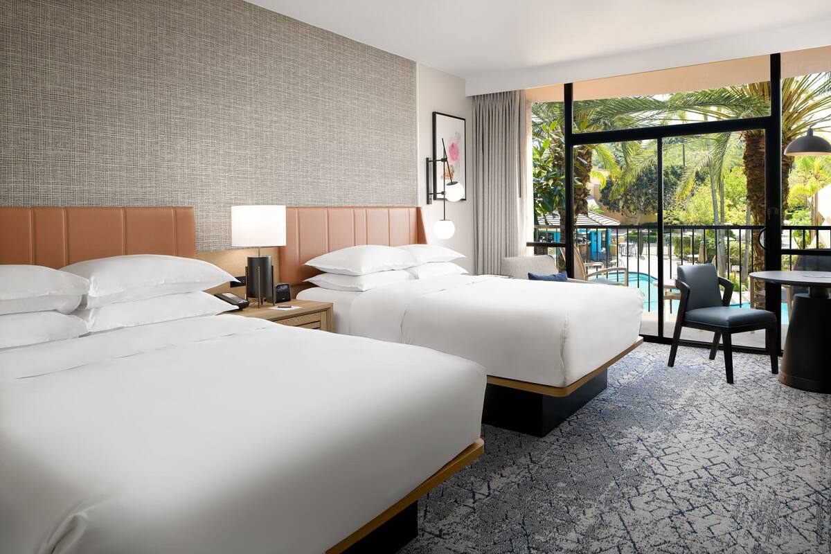 A hotel room with two beds, a desk, a chair, a lamp, and a view of a pool and palm trees.