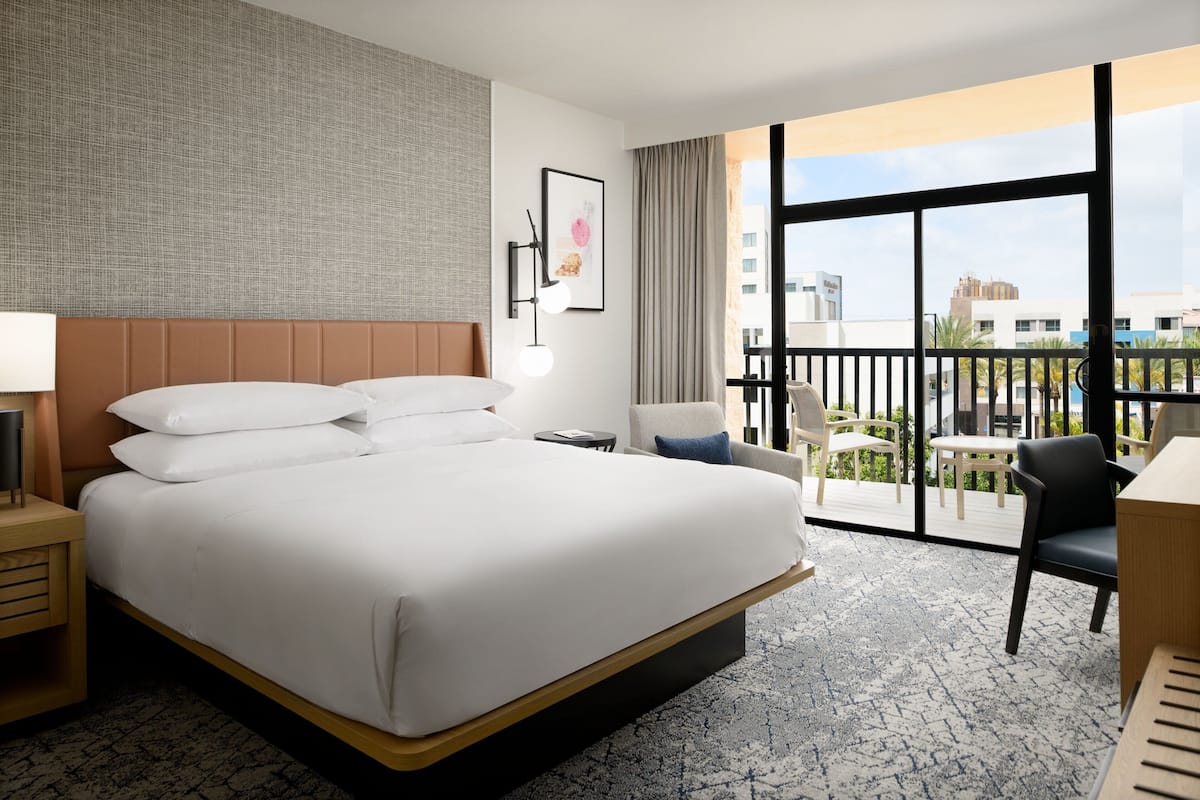 A hotel room with a large bed, a desk, a chair, a small table, a lamp, and a view of the city through large windows.