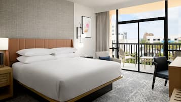 A hotel room with a large bed, a desk, a chair, a small table, a lamp, and a view of the city through large windows.