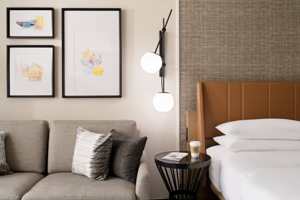 A modern hotel room with a sofa, a round side table with a coffee cup, framed artwork on the wall, and a headboard with a textured fabric.