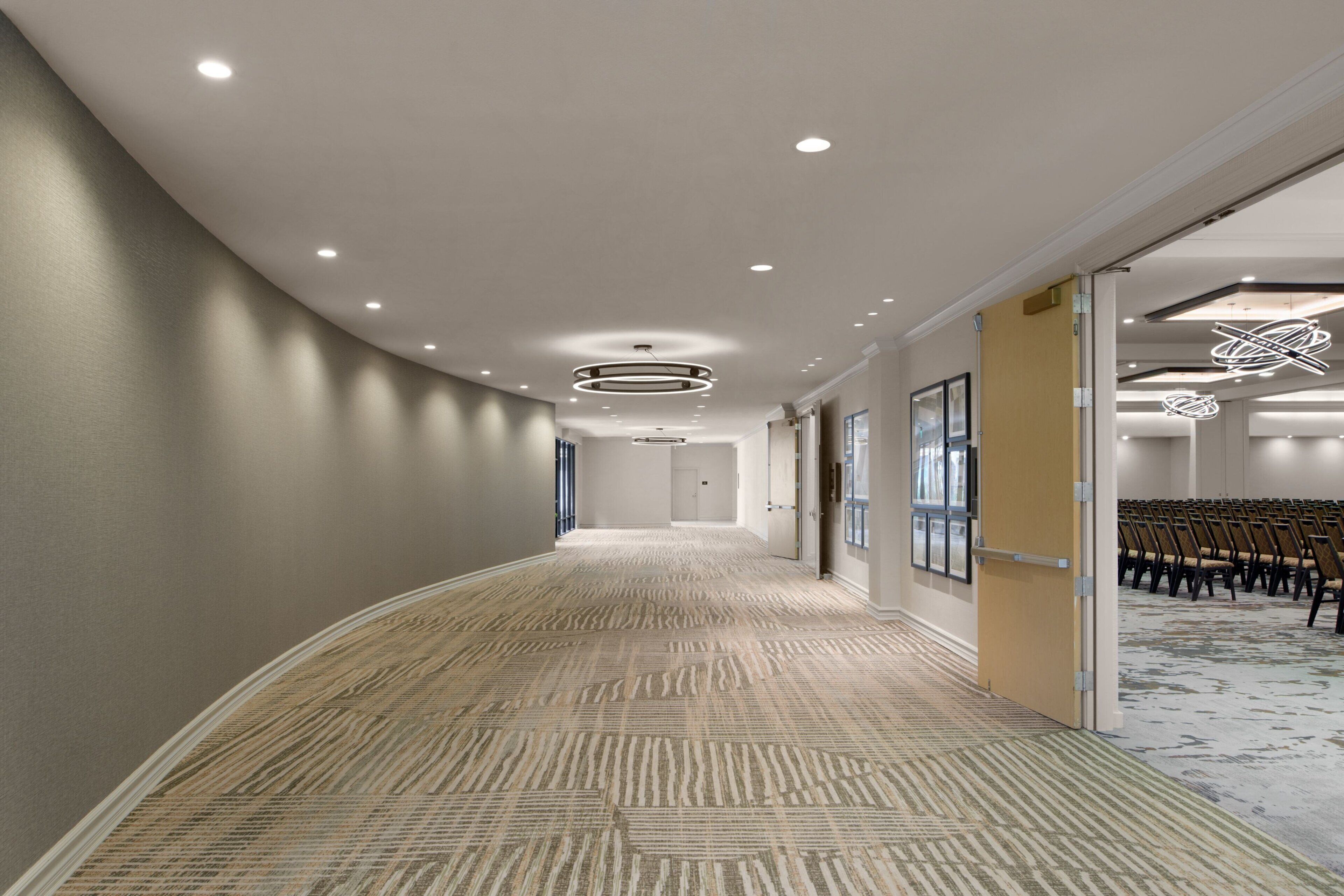 A hotel corridor with a patterned carpet, recessed lighting, and a seating area with chairs.