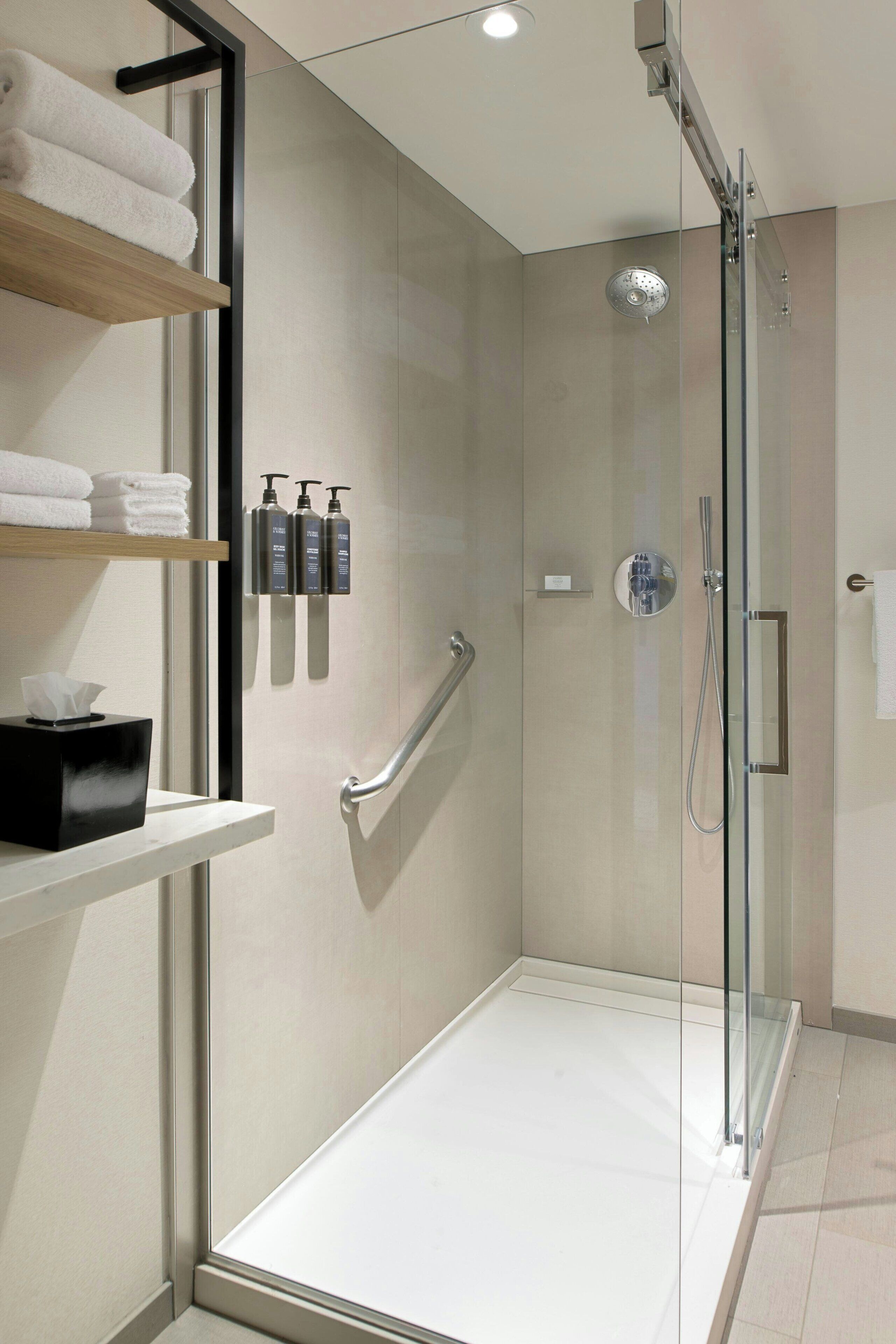 A modern bathroom with a glass shower enclosure, a white sink, and a shelf with towels and toiletries.