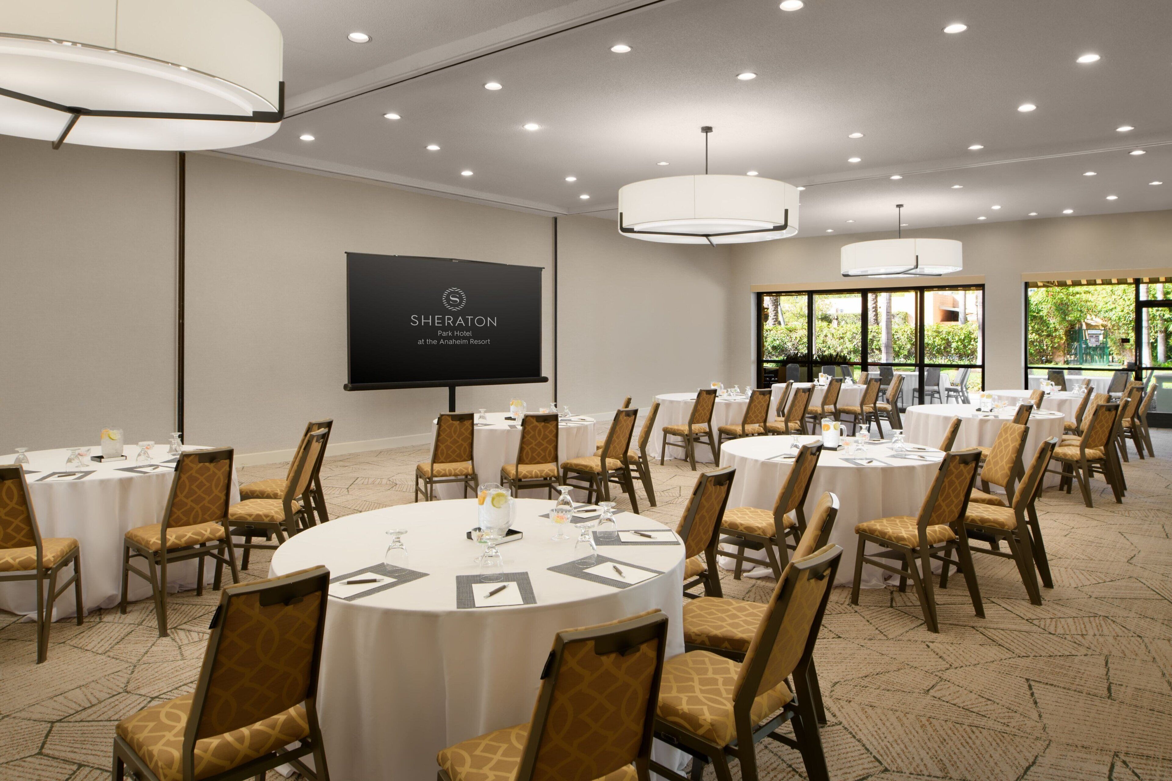 A Sheraton Park Hotel conference room with multiple round tables, chairs, and a large screen displaying the hotel's name.