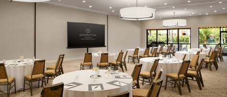 A Sheraton Park Hotel conference room with multiple round tables, chairs, and a large screen displaying the hotel's name.