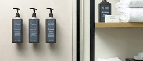 Three bottles of Gilchrist & Soames body wash and conditioner on a wall, a folded white towel on a shelf, and a Gilchrist & Soames skincare bar.