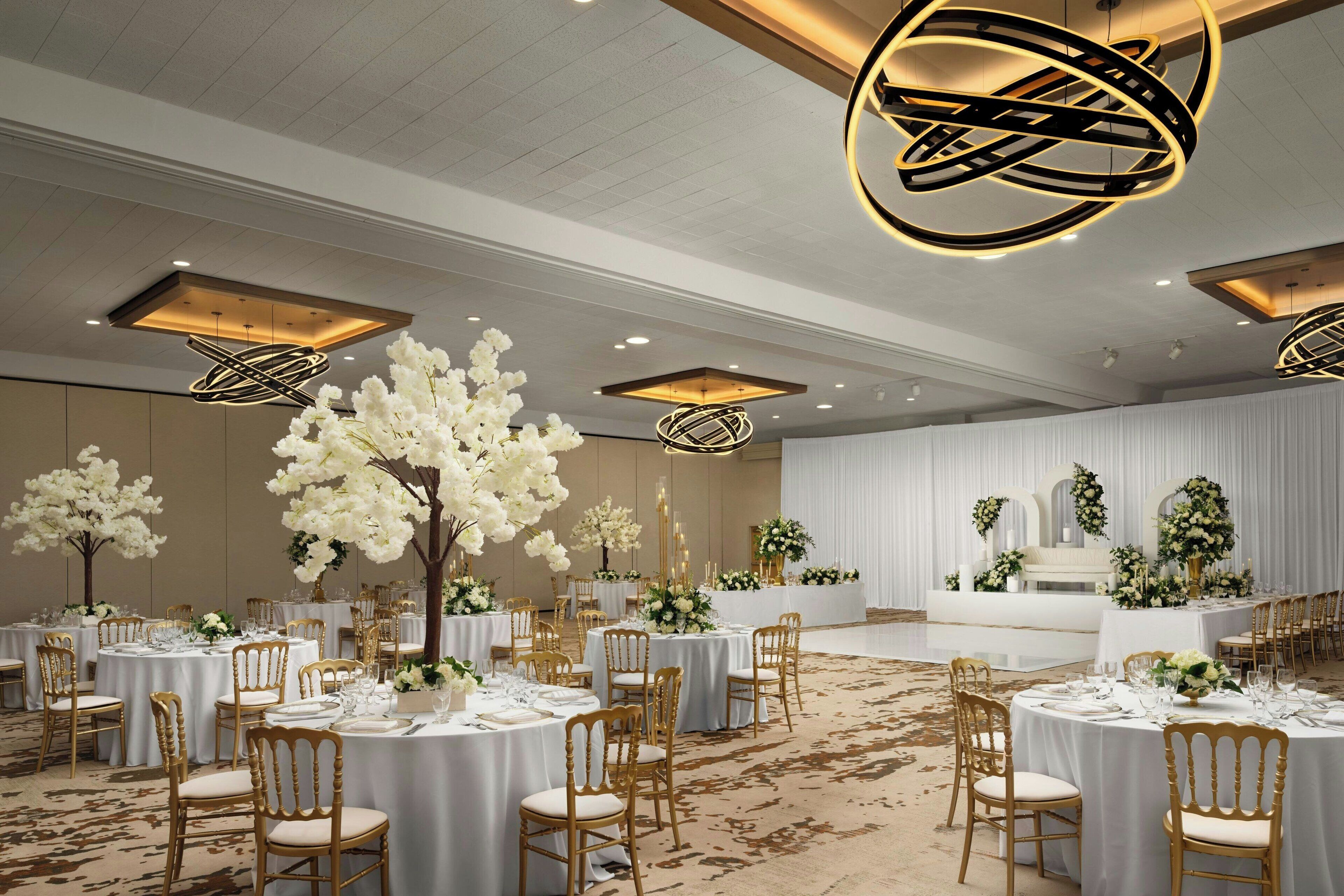 A banquet hall with round tables, gold chairs, and white floral arrangements.