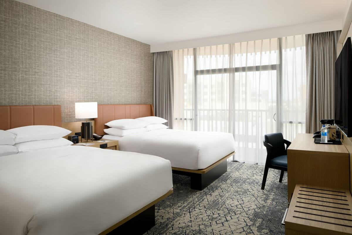 A hotel room with two beds, a desk, a chair, and a large window with curtains.