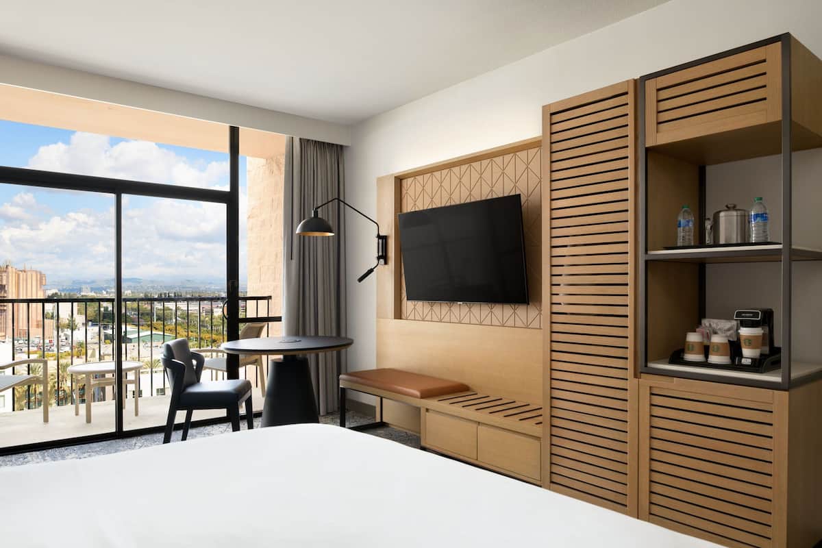 A hotel room with a large window, a bed, a desk, a chair, a bench, a TV, and a shelf with coffee cups.