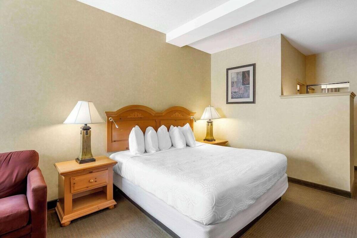 A hotel room with a large bed, two bedside tables with lamps, a wooden headboard, a single chair, and a framed picture on the wall.