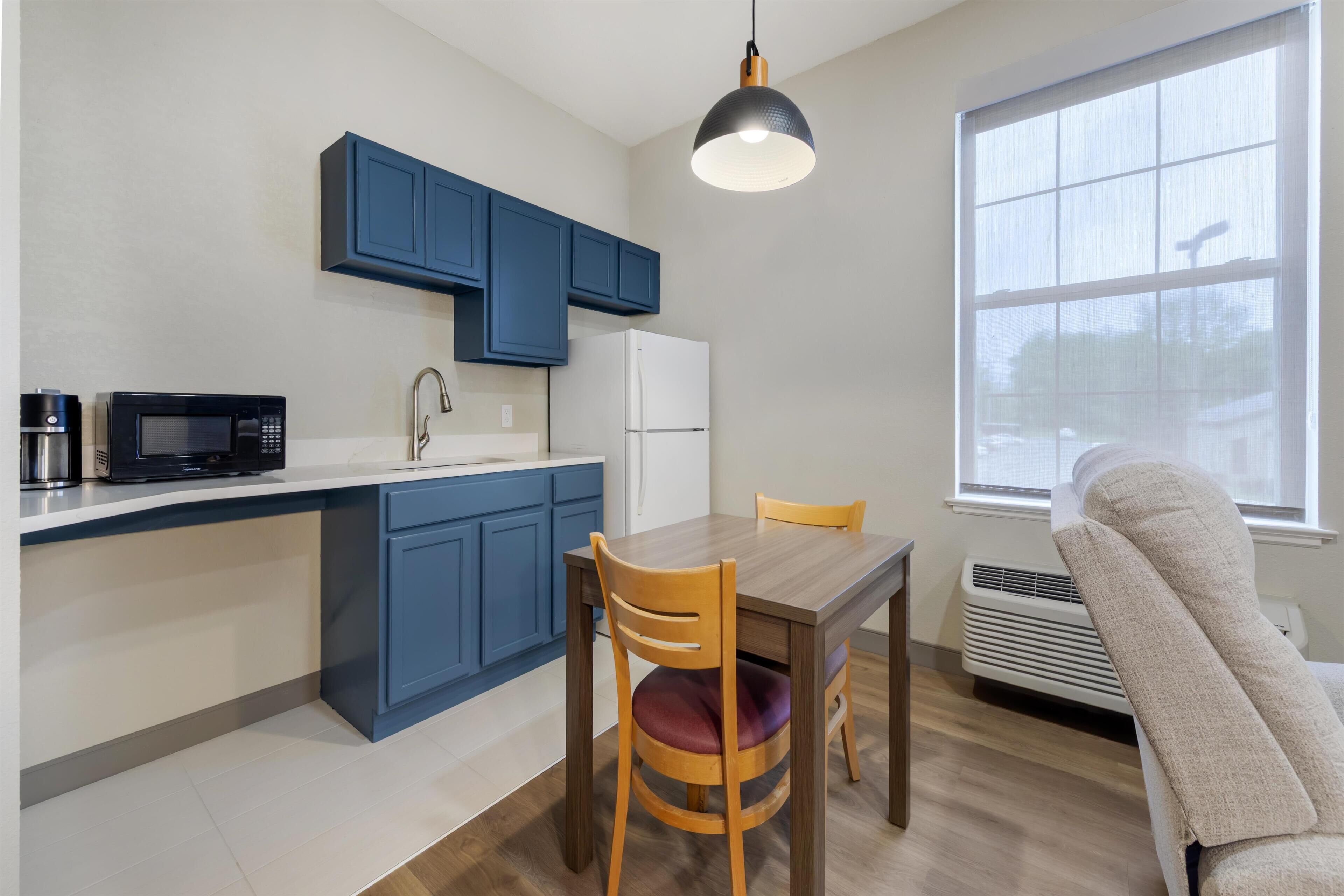 A compact kitchen with blue cabinets, a microwave, a refrigerator, a dining table with chairs, and a large window.