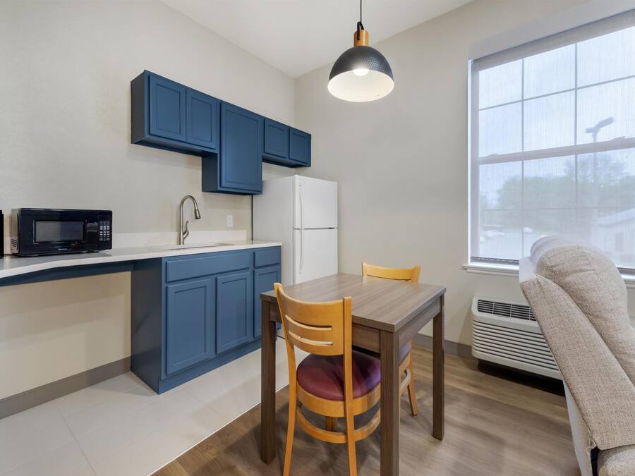 A compact kitchen with blue cabinets, a microwave, a refrigerator, a dining table with chairs, and a large window.