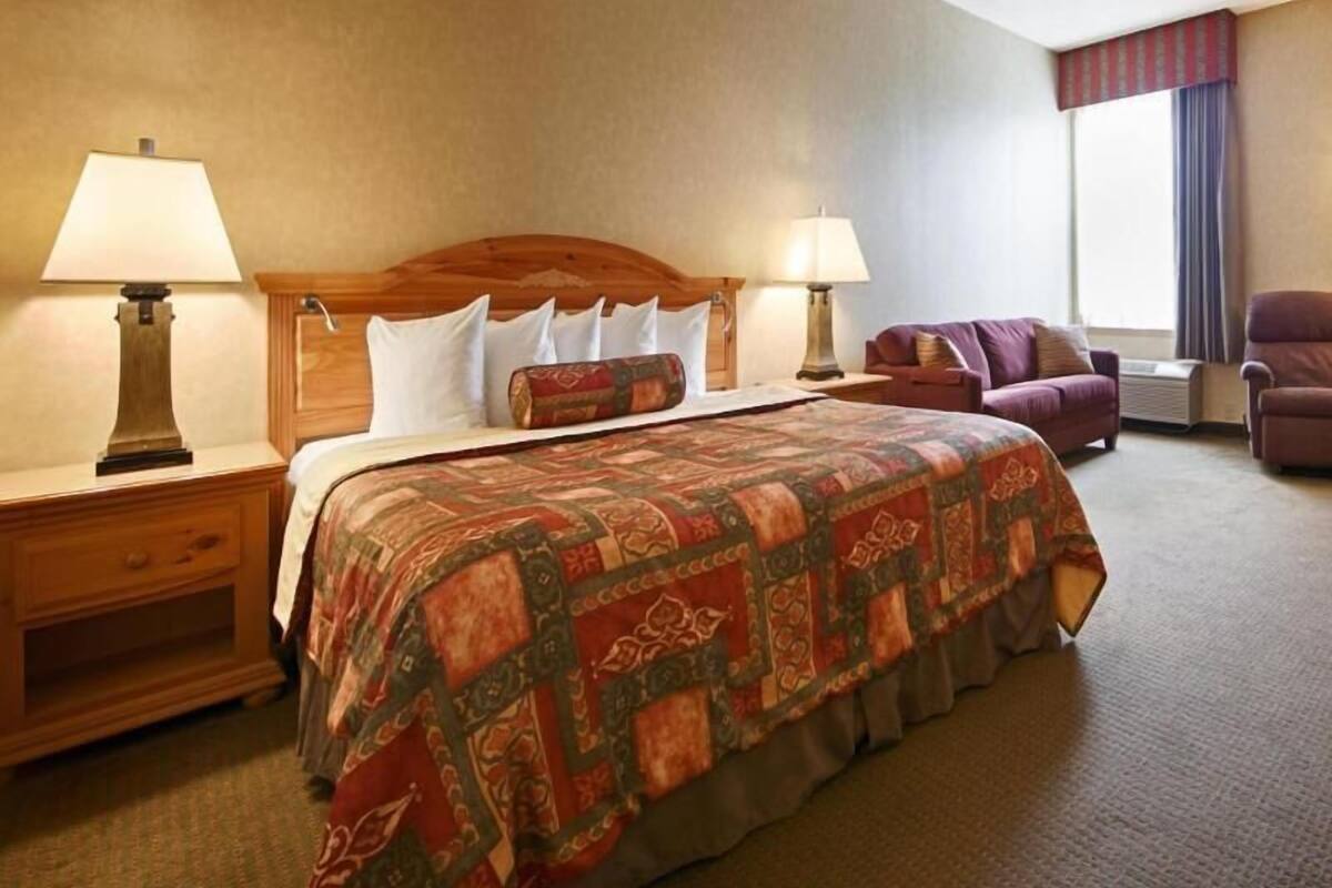 A hotel room with a large bed, two bedside tables with lamps, a wooden headboard, a purple armchair, and a window with curtains.