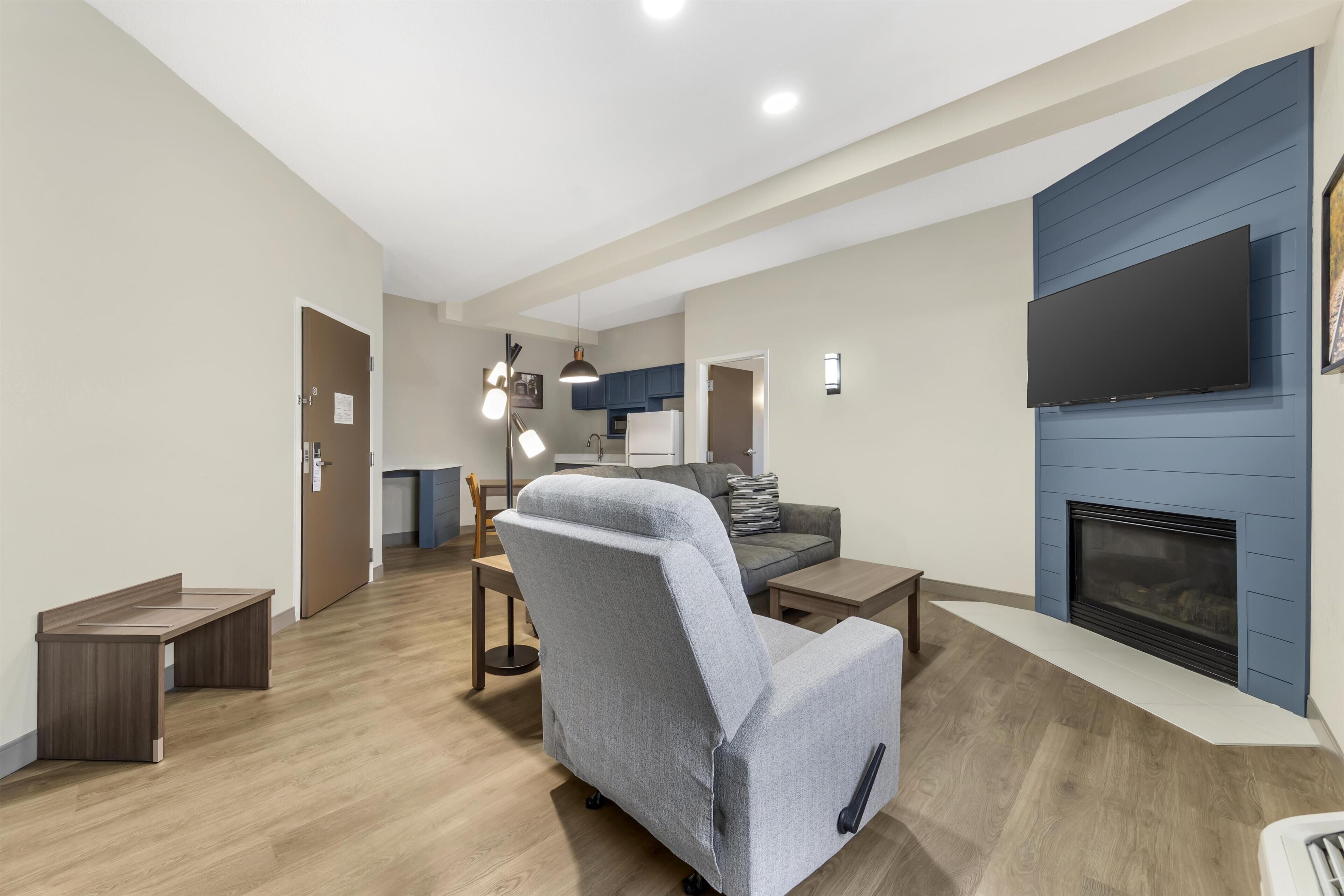 A modern living room with a grey armchair, a flat-screen TV mounted on a blue wall, a fireplace, and a wooden floor.