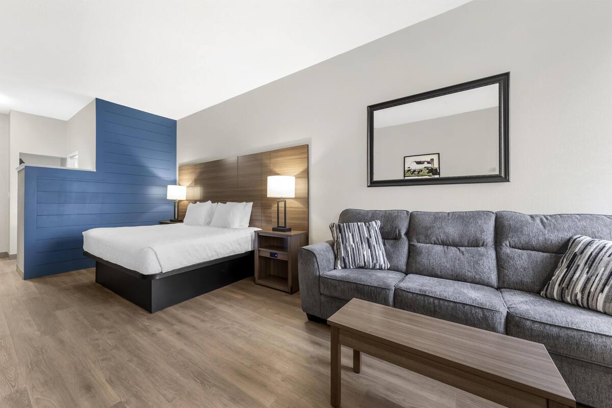 A modern hotel room with a bed, a gray sofa, a wooden coffee table, a nightstand, and a wall-mounted mirror.