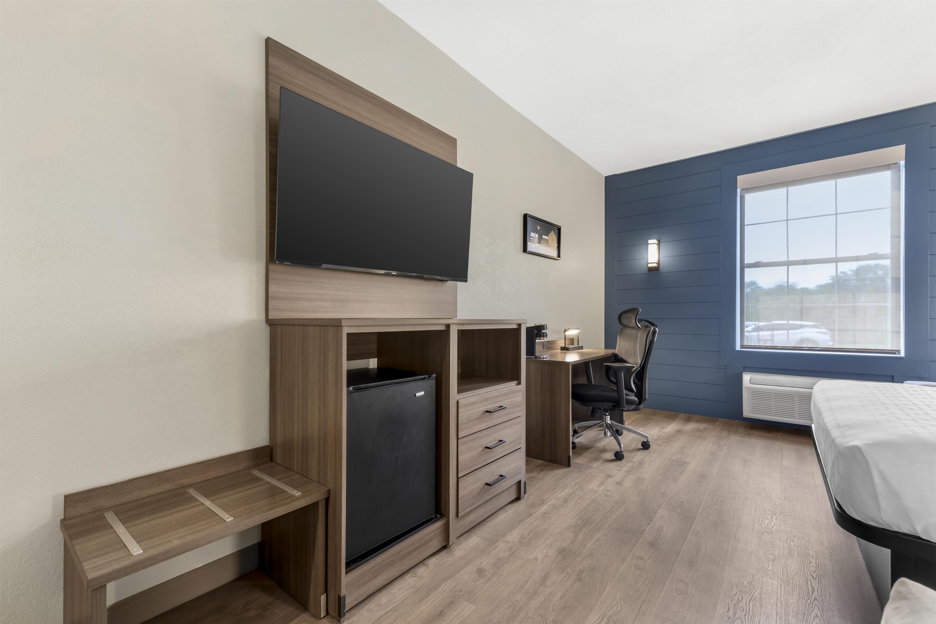 Quarto com cama, escrivaninha, cadeira, televisão e um pequeno frigorífico.